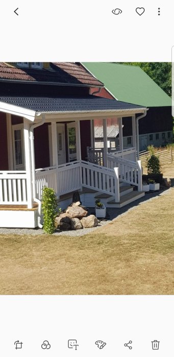 Nybyggd trädäck och altan i vitt med räcken vid ett rött hus med grönt tak under en klarblå himmel.