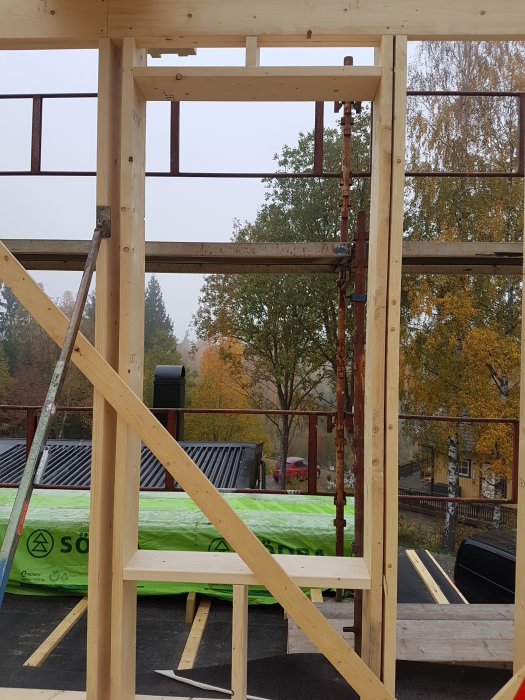 Trästomme för andra våningen i byggprojekt, med takreglar och fönster upplyfta, omgivet av höstlövsträd.