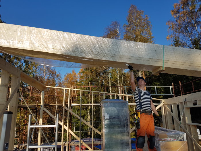 Person i byggarbetskläder lyfter stort träregel på byggnadsställning mot klarblå himmel.