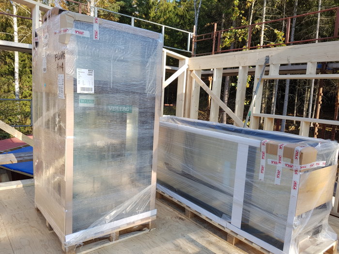 Förpackade takreglar och fönster på en byggarbetsplats med trästomme och skog i bakgrunden.