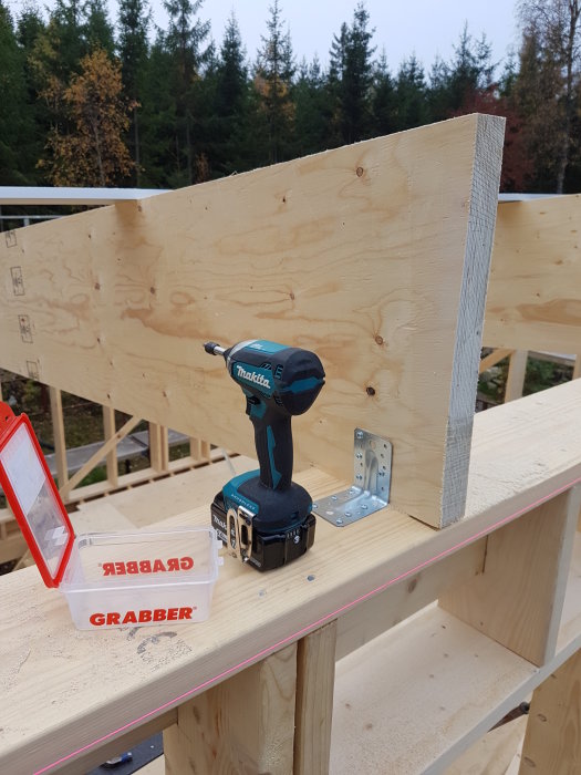 Verktyg och skruvar på en träram i byggprocessen med synliga skruvreglar och bakgrunden av träd.