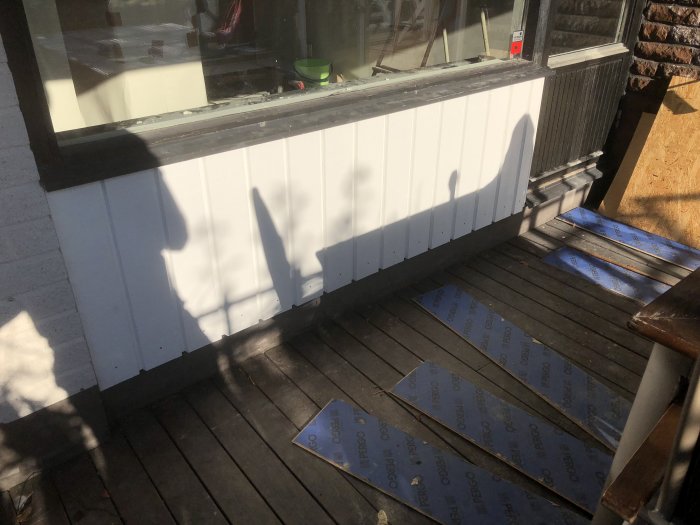 Vitt täckstycke på en husvägg ovanför altanens trägolv, skuggor syns på panelen och oisolerad isolering på golvet.