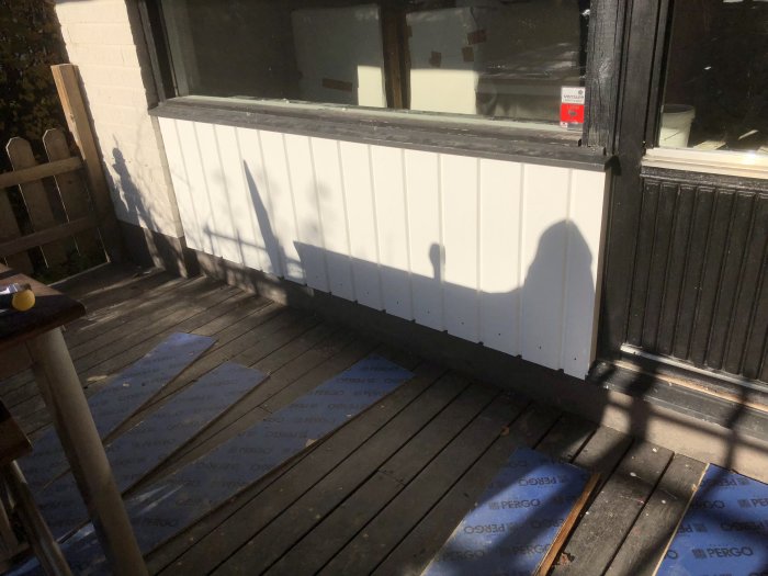 Vit panel på en altan med skuggor på golvet och en del förpackningsmaterial för golv kvar på trätrallen.