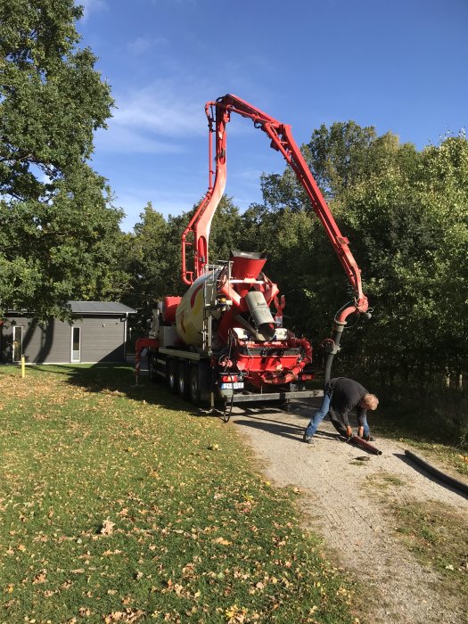 Gjutbil med röd arm och slangar för betong, och en person som arbetar med ett PVC-rör nära hus.