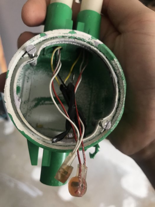 Hand som håller en grön cirkulär elektrisk dosa med flera oanslutna röda, gula, vita och svarta kablar som sticker ut.