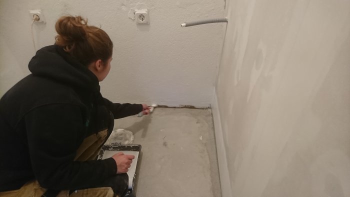 Person applicerar våtrumsspackel längs golv mot vägg i pågående renovering av tvättstuga.