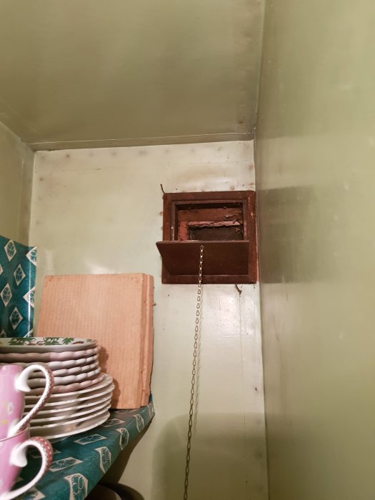 Gammalt skafferi med öppen ventilation i väggen och en dragningskedja, omgiven av grönmålade väggar och tallrikar på hylla.