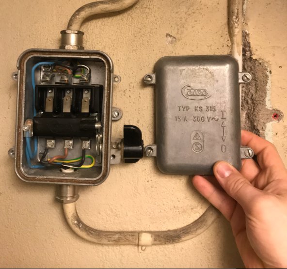 Hand håller en gammal ASEA Cewe KS315 säkringslåda med öppet lock som visar säkringar och ledningar på en vägg.