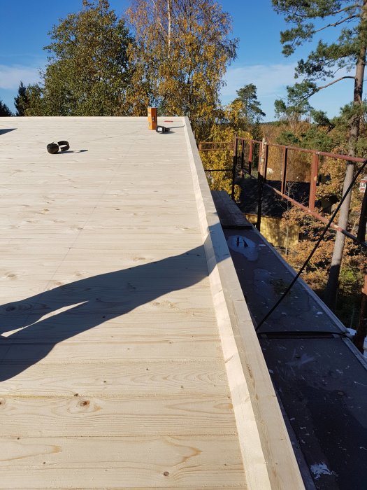 Obehandlat träunderlag på ett tak med en konstruktionsnivå, en rullad takpapp vid kanten och en vattenpass i bakgrunden, klar himmel.