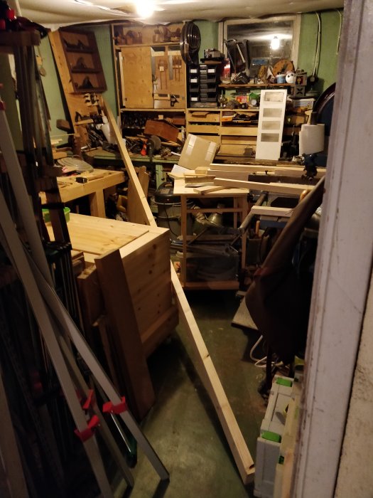 En verkstad överbelamrad med träplankor och verktyg, inklusive en stor träbit som lutar mot en arbetsbänk.