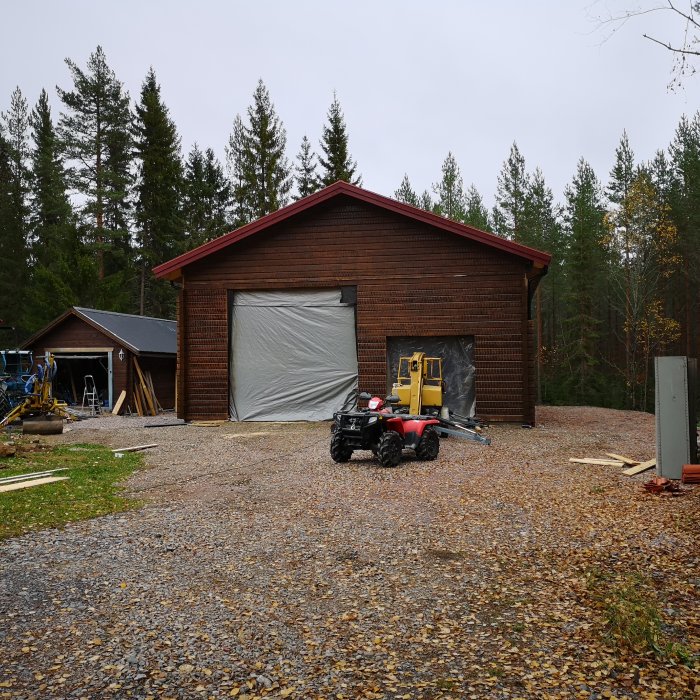 En rödbrun tegelgaragebyggnad under konstruktion med högt tak, omgiven av träd och med en fyrhjuling framför.