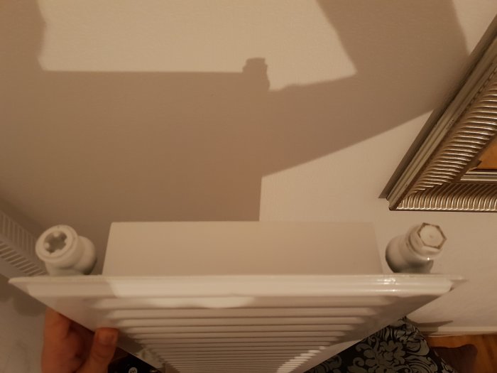 Hand håller upp en vit radiator med ovanligt formade ventiler i ett rum.