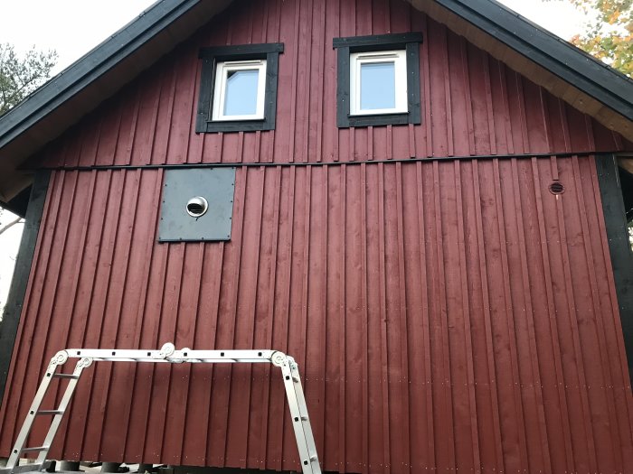 Röd plåtfasad på hus med utblås, två fönster och stege framför.