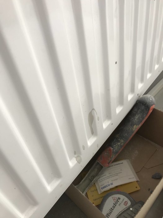 Otäckt vitmålad radiator med stänk av färg och en synlig kvist, intill en låda med byggmaterial.