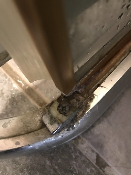 Trasig hållare vid foten av en duschvägg med synlig rost och skador på glasets infästning.