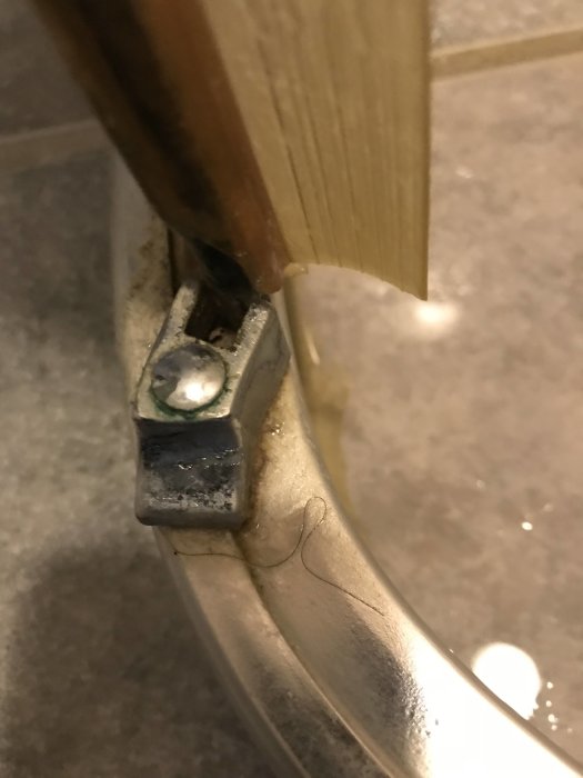 Närbild på en trasig metallhållare i nederkant av en duschvägg, vid kakel och duschdörr.