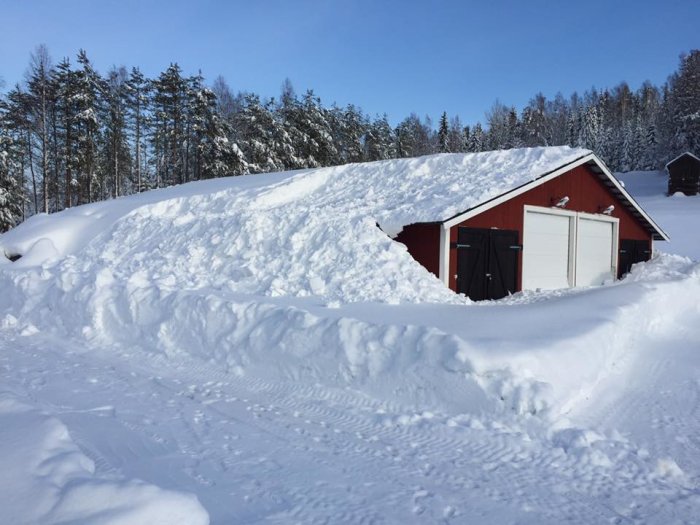 Rött hus delvis täckt av tjockt lager snö med solpaneler på taket och omringat av träd.