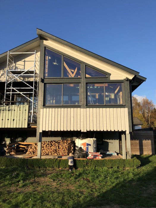 Husfasad med stora fönsterpartier och nyinstallerat stuprör, utelampa och ved under byggställning.