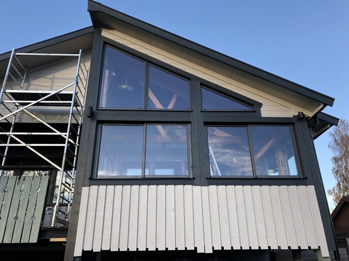 Husfasad med stora fönster, nyligen monterade stuprör och utelampa, samt nedtagen byggställning.