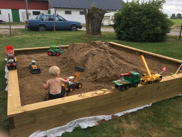 Barn leker i en sandlåda med leksaksbilar och lastmaskiner, inramad av träplank, utomhus.