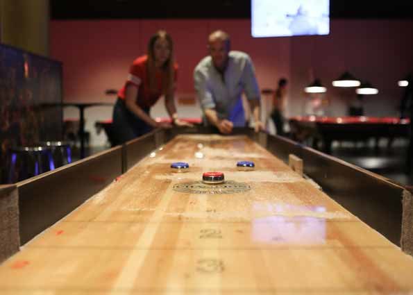 Två personer spelar shuffleboard vid ett långt, stavlimmat träbord.