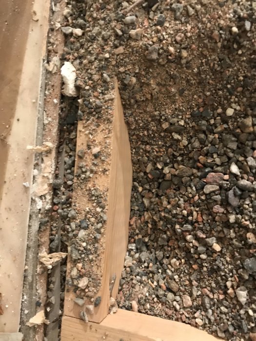 Hål i golvet med synliga träreglar, sand och grus, omgivet av intakt golvspån och plast.
