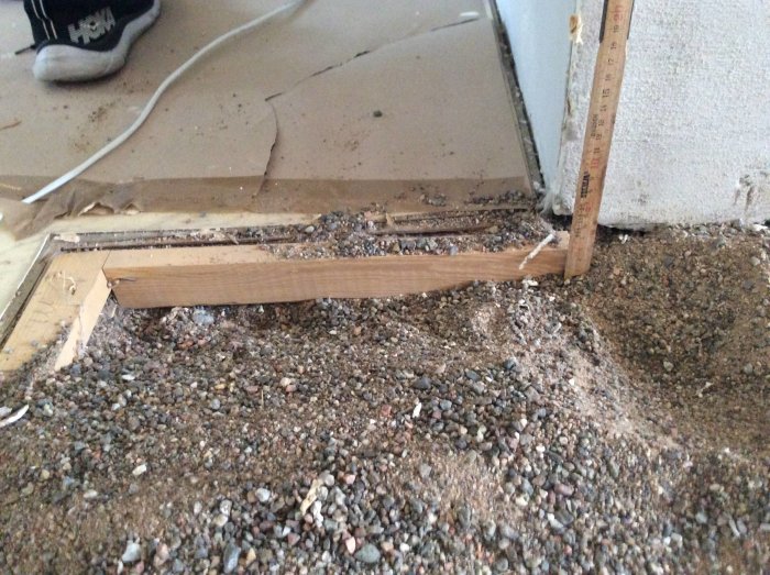 Upprivet golv med synliga träreglar och ett lager av sand, linjal visar tjocklek; skadat golvspån och lumppapp syns i kanten.