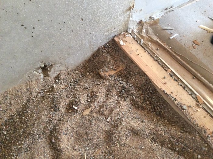 Hörn av ett rum med upprivet golv som visar sand, spånskivor och delvis dolda träreglar.