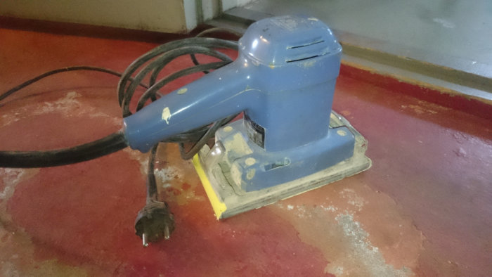Gammaldags blå slipmaskin i gjutjärn på ett slitet rött och betongfärgat golv.