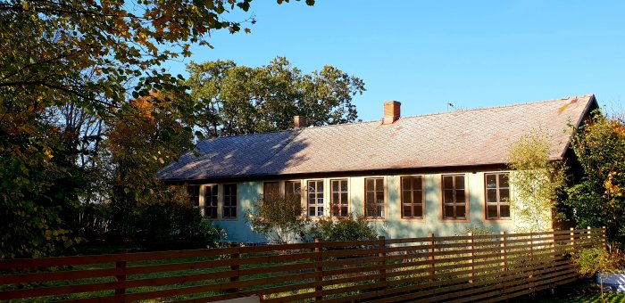 En gammal skolbyggnad med blåa väggar och brun tegeltak omgiven av träd och en trästaket i solljus.