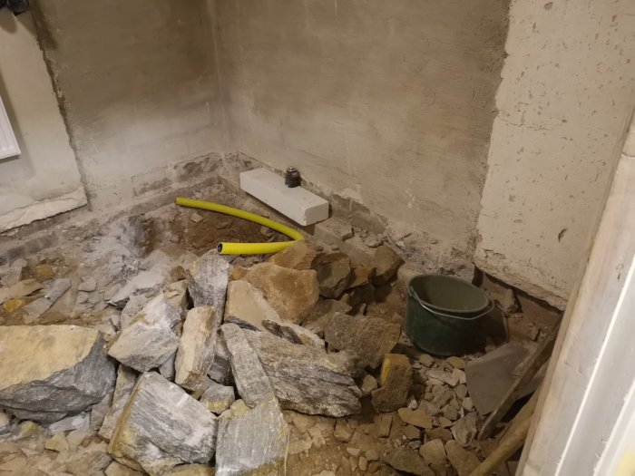 En källarhörna under renovering med berg och grus synligt efter grävningsarbete, omgiven av källarväggar och verktyg.