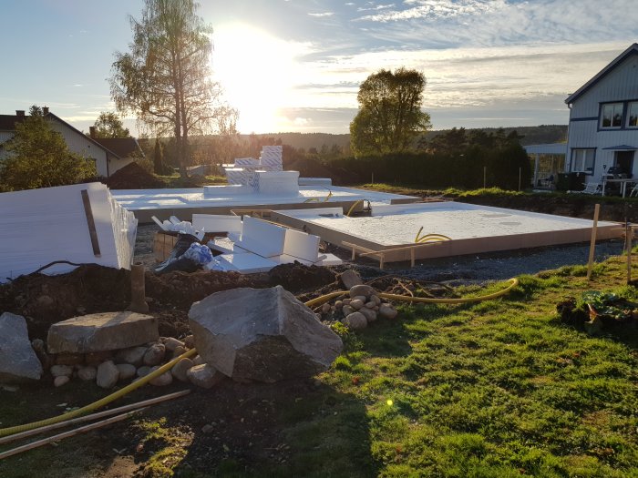 Grundläggning för hus med isolerplattor och armeringsjärn framför grävd kulvert i ett bostadsområde.