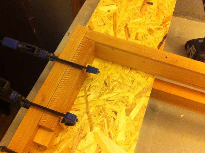Konstruktion av en träspaljé där två trästycken hålls samman med klämmor på en OSB-skiva på ett verkstadsgolv.