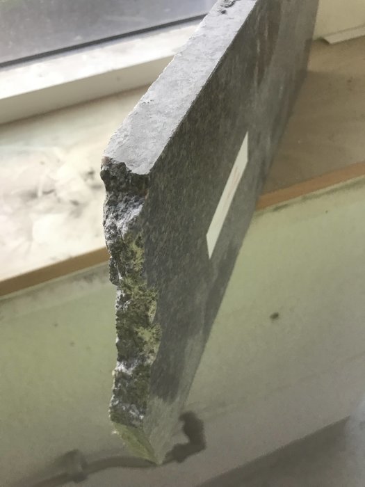 Kant av ett granitblock som visar dess struktur och färg, med bakgrund av ett fönster och golv.