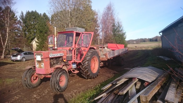 Röd Volvo BM 650 traktor parkerad utomhus med släpvagn, omgiven av skog och lantlig miljö.