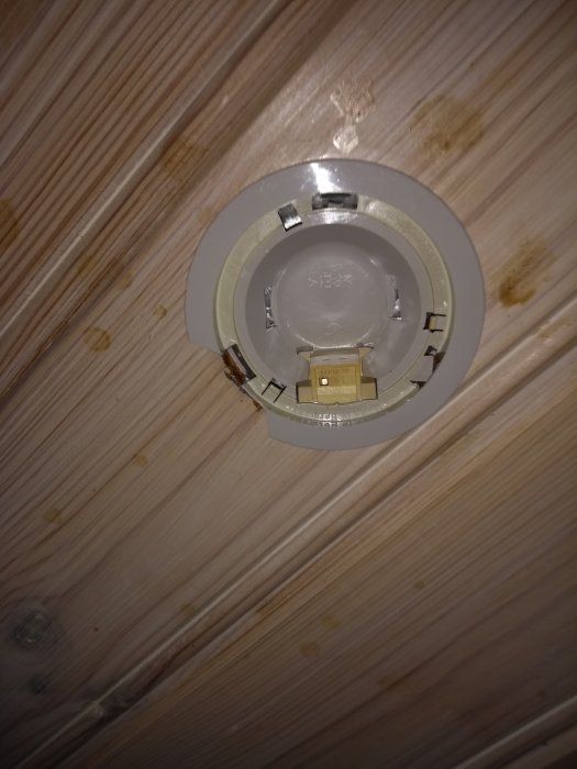 Skadad downlight i taket som behöver tas bort, omgiven av träpanel, med synliga tecken på slitage och åldring.