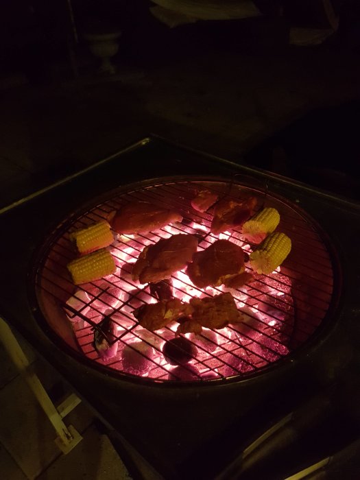 Kolgrill i mörker med glödande kol, grillat kött och majs, i ett halvfärdigt utekök.