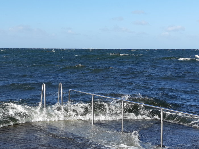 Bild på upprörda vatten med vågor som slår mot en badstege under blåsig himmel.