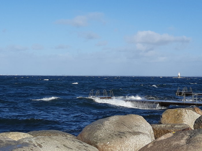 Havsvy med vågor och vind, brygga delvis täckt av vatten, och fyren i bakgrunden.
