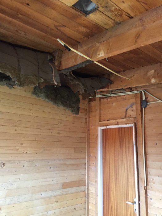 Rivet innertak med synliga träbjälkar och isolering ovanför en träpanelvägg och en brun dörr.