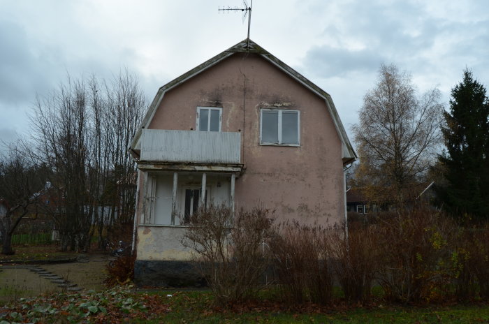 Äldre rosa husfasad med behov av renovering, en balkong, slitet tak och en övergiven trädgård.