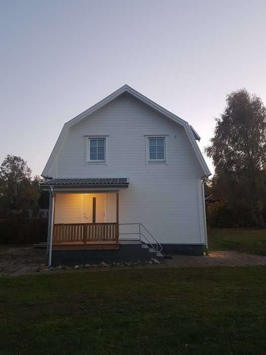 Tvåvånings hus med vit fasad, veranda och gräsmatta i skymningen.