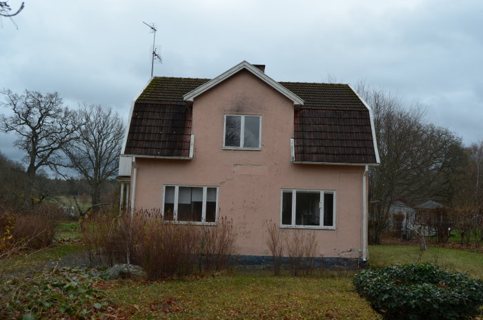 Rosa hus med slitet tegeltak och synliga mossbefläckningar, trädgård med övervuxen vegetation.