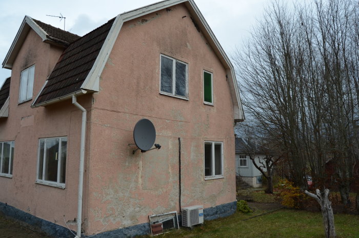 Slitet rosa tvåvåningshus med flagnande fasad, satellitantenn och omgivande trädgård.