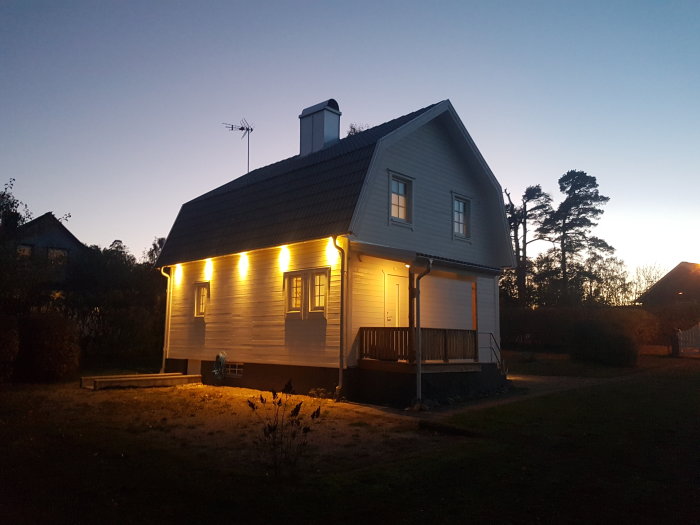 Litet hus i kvällsljus med belysning, ej renoverat exteriört, med antenner på taket.