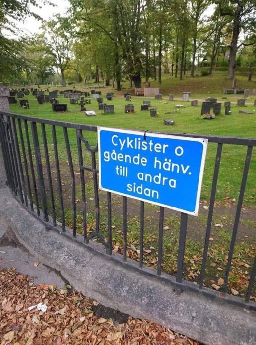 Skylt "Cyklister o gående hänv. till andra sidan" framför kyrkogård med gravstenar och träd.