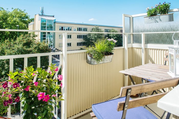 Solig balkong med träbord, stolar och blomlådor mot bakgrund av stadsmiljö.