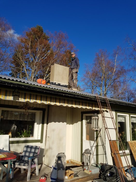 En person på taket arbetar på en ofärdig skorsten med verktyg och byggmaterial runtomkring.
