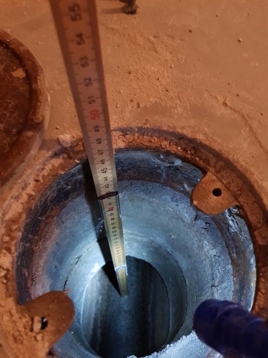 Måttband som visar djupet av ett hål till en stamledning i en betongplatta med ett lock intill.