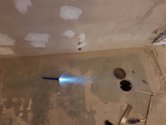 Bild av ett betonggolv i en källare med föreslagen plats för ny golvbrunn markerad med en ficklampa och befintligt hål med lock.